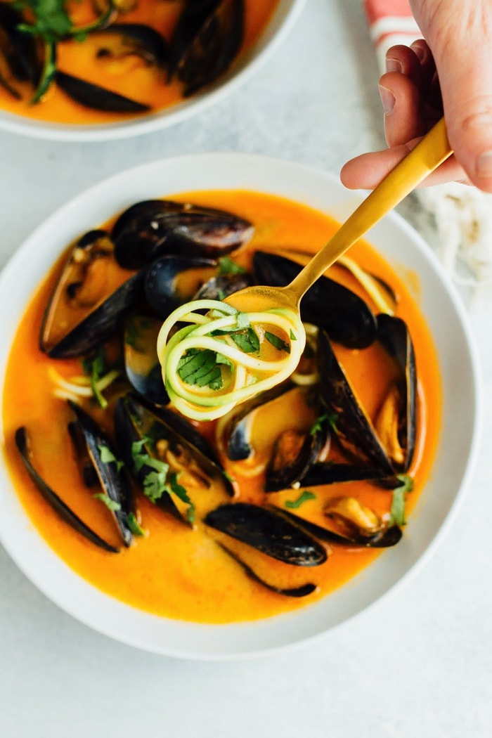 EASY COCONUT CURRY MUSSELS WITH ZUCCHINI NOODLES // paleo and gluten-free Hand holding a spoon of the coconut curry broth with zucchini noodles. Bowl of mussels in the background.