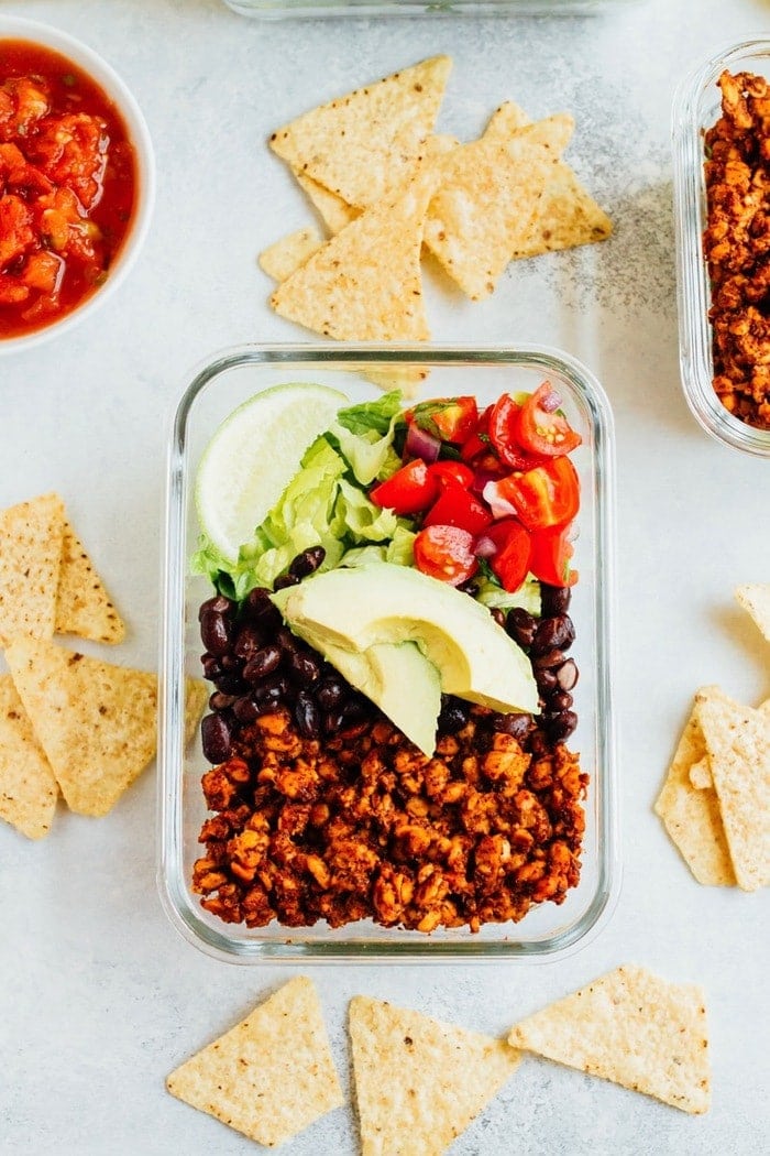 Tempeh Taco Salad Meal Prep Bowls with tempeh taco meat, black beans, tomatoes, onions and avocado A glass container with tempeh, black beans, cherry tomatoes, lime, and lettuce.