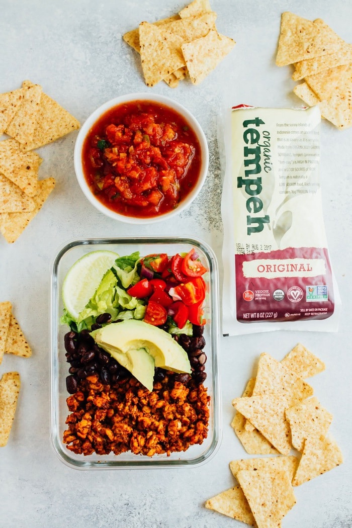 Tempeh Taco Salad Meal Prep Bowls with tempeh taco meat, black beans, tomatoes, onions and avocado A glass container with seasoned and cooked tempeh, beans, tomatoes, avocado, and lettuce. Tortilla chips, a bowl of salsa, and a packet of organic tempeh are on each side of the container.