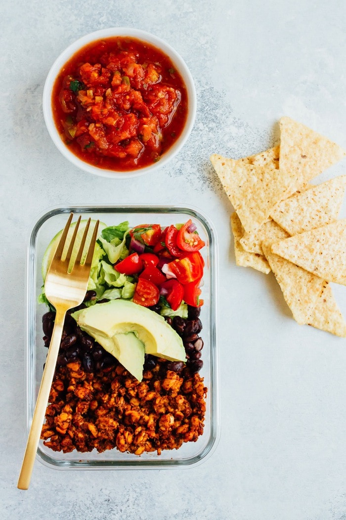 Tempeh Taco Salad Meal Prep Bowls with tempeh taco meat, black beans, tomatoes, onions and avocado A container of a tempeh taco salad with tortilla chips and a small bowl of salsa to the side.