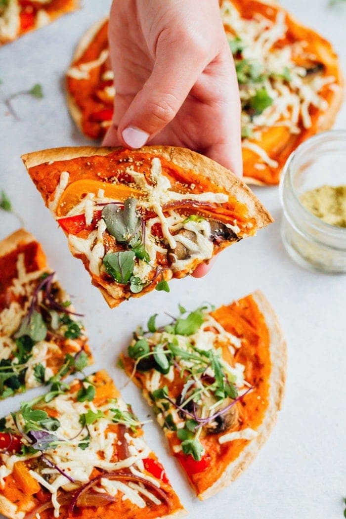 Get creative on pizza night with these thin and crispy vegan tortilla pizzas. They're easy, personal-size (for instant portion control) and delicious! Hand holding a slice fo tortilla hummus pizza topped with vegan cheese and veggies.