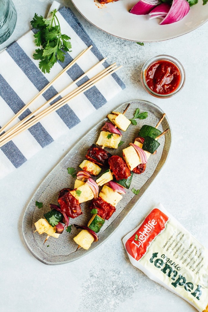 Bring on the BBQ tempeh skewers! You’ll love this plant-based grilling recipe where nutty tempeh is coated in homemade BBQ sauce and grilled to perfection with zucchini, summer squash and red onion. Three bbq tempeh skewers with onion, pineapple and zucchini on a plate. A small glass container of BBQ is to the side as well as a packet of tempeh.