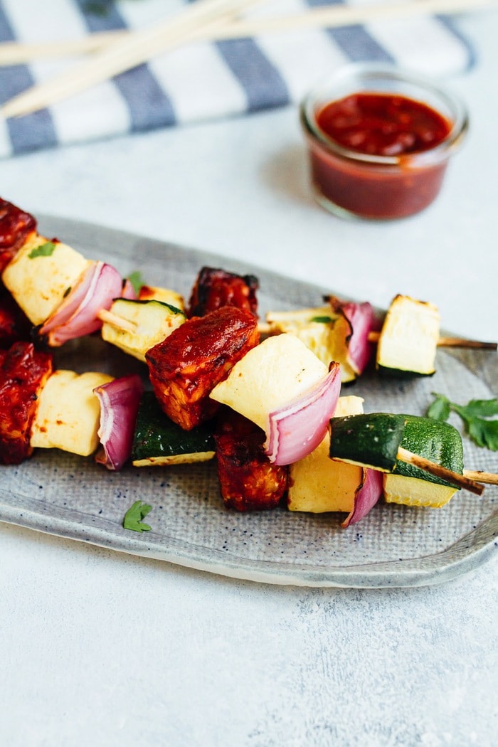 Bring on the BBQ tempeh skewers! You’ll love this plant-based grilling recipe where nutty tempeh is coated in homemade BBQ sauce and grilled to perfection with zucchini, summer squash and red onion. Three bbq tempeh skewers served on a plate with a glass container of BBQ to the side.