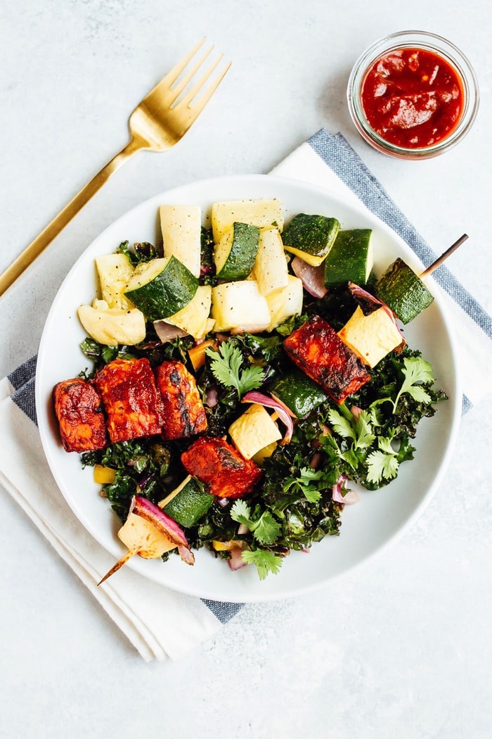 BBQ TEMPEH SKEWER SALAD // You’ll love this plant-based grilling recipe where nutty tempeh is coated in homemade BBQ sauce and grilled to perfection with zucchini, summer squash and red onion. Bbq tempeh skewers with onion, pineapple, and zucchini on a salad with a dish of bbq sauce next to the plate.