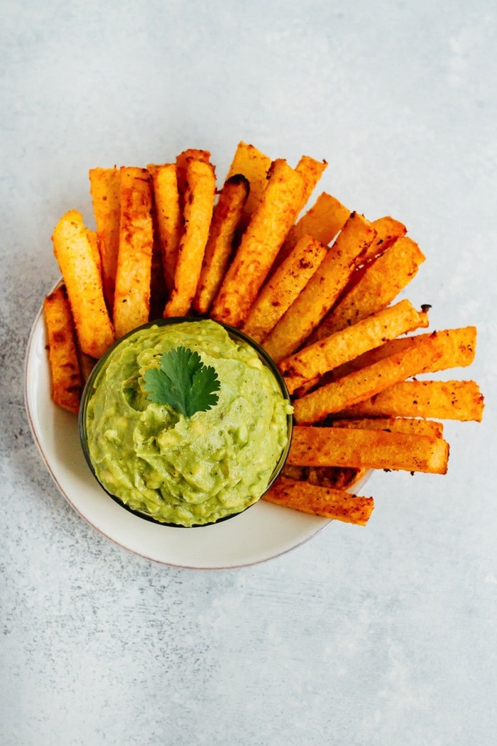 Golden Gut Baked Jicama Fries with Turmeric and Black Pepper. Serve with fresh guacamole for a delicious summer side. A serving of crunchy jicama fries with a small bowl of guacamole in the center of the bowl.