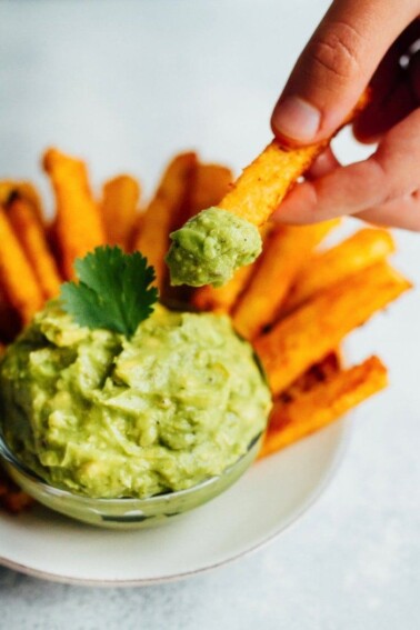 A hand holds a jicama fry that has been dipped into a small bowl of guacamole.