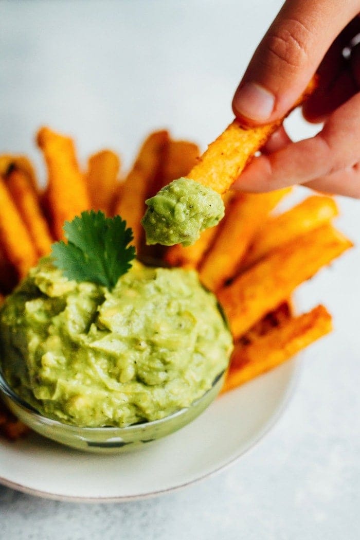 Golden Gut Baked Jicama Fries with Turmeric and Black Pepper. Serve with fresh guacamole for a delicious summer side. A hand holds a jicama fry that has been dipped into a small bowl of guacamole.