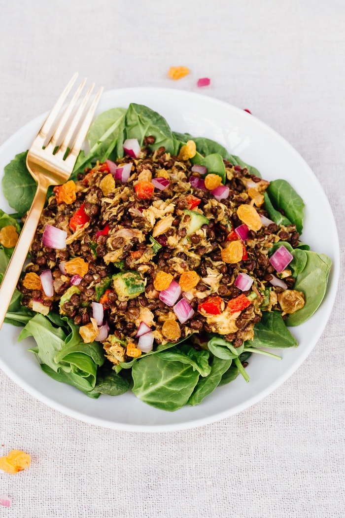 This lentil tuna salad combines beluga lentils with canned tuna, fresh veggies and a turmeric dressing. It's absolutely delicious and perfect for sharing at summer cookouts and parties. A serving of lentil tuna salad on a bed of fresh greens on a white plate with a metal fork to the side.