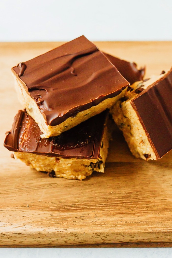 No Bake VEGAN GLUTEN-FREE Cookie Dough Bars Multiple cookie dough bars stacked on one another on a wooden surface. The bars have a chocolate coating on top.