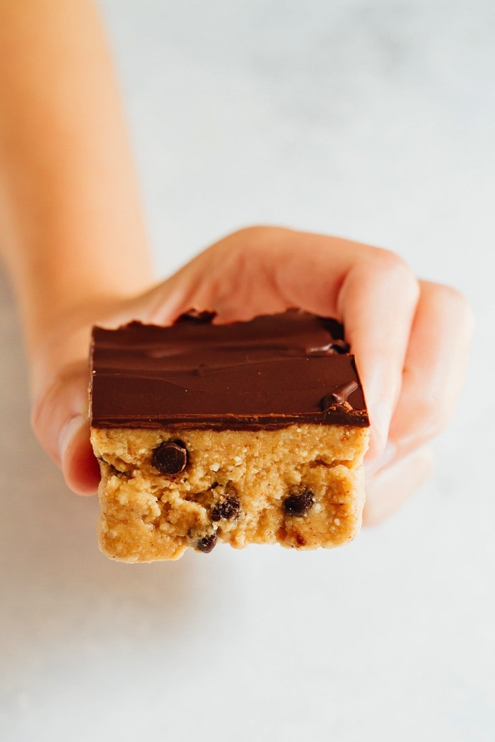 No Bake VEGAN GLUTEN-FREE Cookie Dough Bars A womans hand hold a cookie dough bar with a chocolate coating on top.