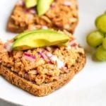 A serving of tuna salad on two slices of bread topped with slice avocado and green grapes on the side.
