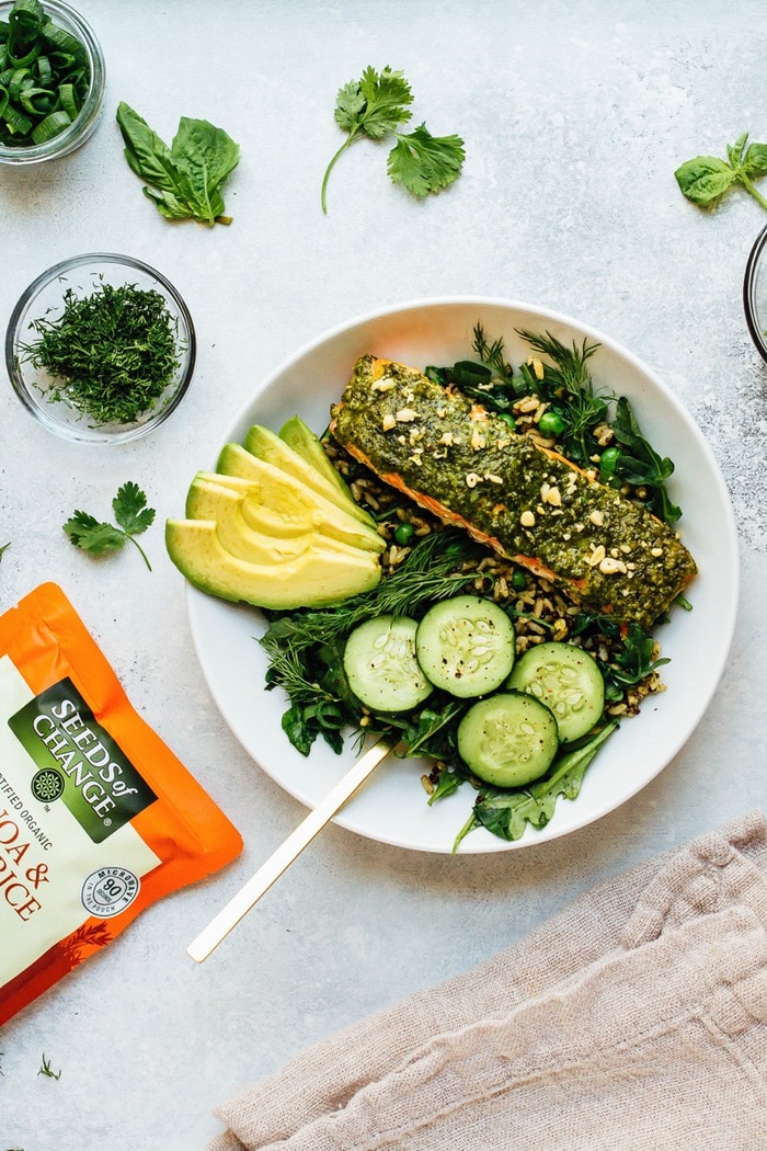 Green Goodness Bowl with Baked Steelhead Trout Pesto salmon, greens, herbs, cucumber, and avocado over rice, on a white plate surrounded by bowls of herbs.