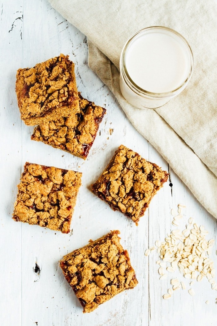 Peanut Butter and Jelly Crumble Bars Peanut Butter and Jelly Crumble Bars on a table next to some oats and a glass of almond milk.