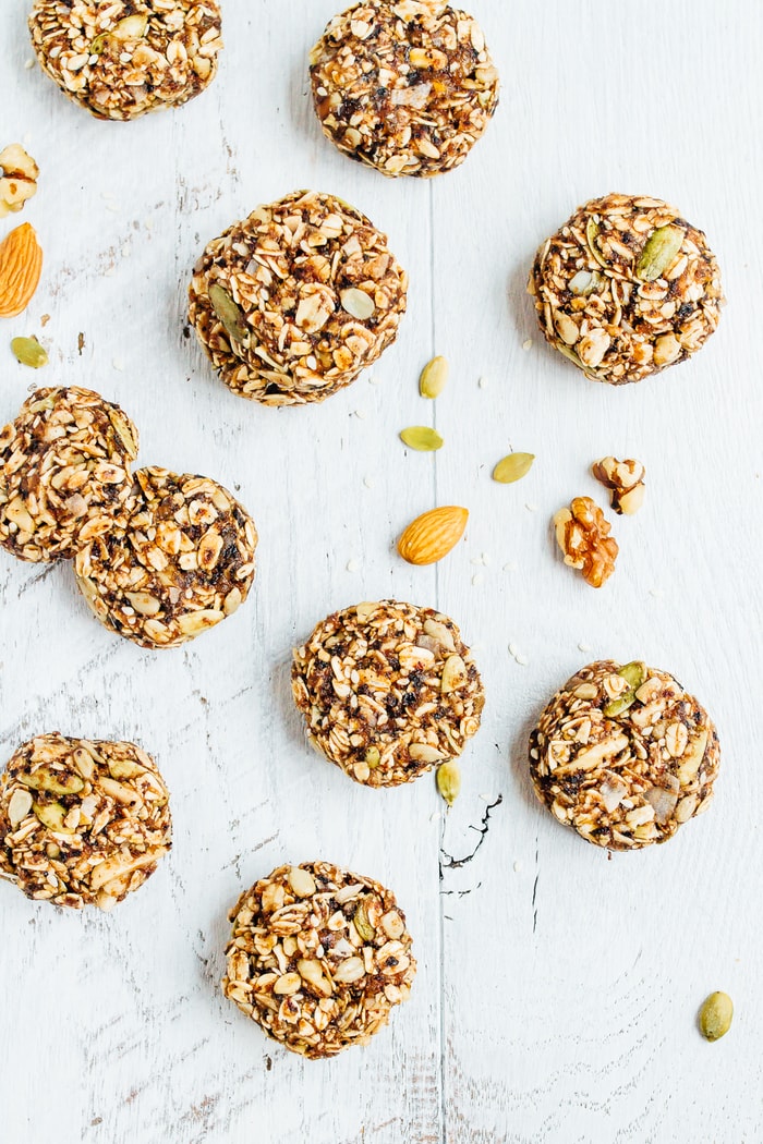 Eat like a bird with these BIRD FOOD ENERGY BITES! Made with whole grain oats, nuts, seeds and dried fruit, these bites are a nutrient-rich treat perfect for on-the-go snacking. Vegan + gluten-free. Granola bites on a white wooden surface, surrounded by seeds and nuts.