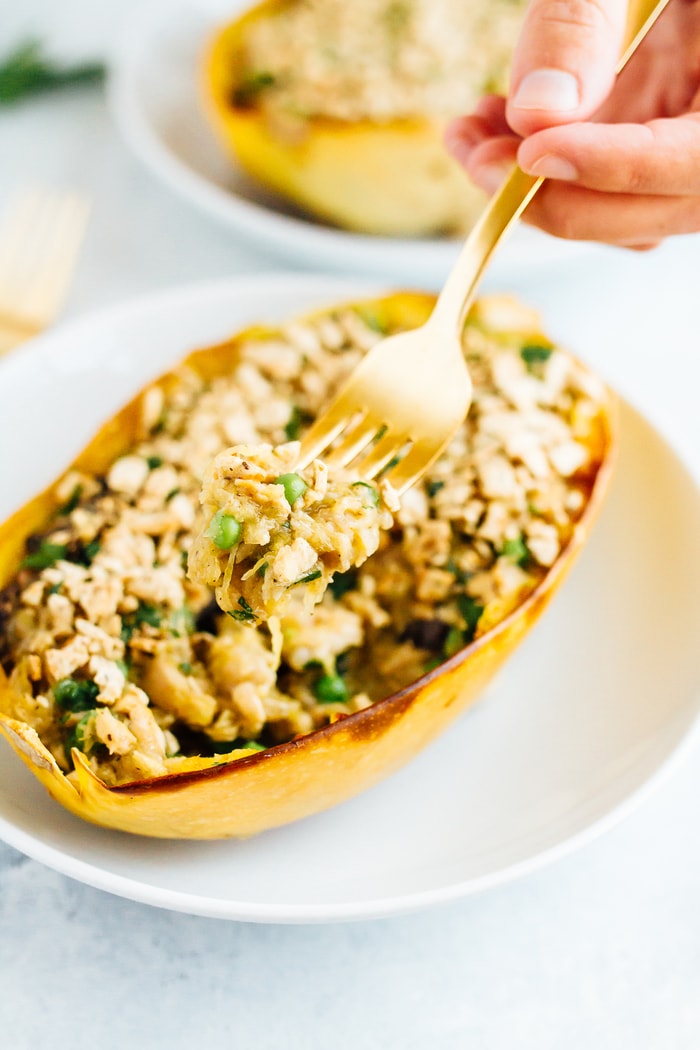 A healthy spin on a comfort food classic, these spaghetti squash tuna noodle casserole boats are filled with loads of creamy noodle goodness. Gluten-free, dairy-free, low-carb and paleo! A hand scooping with a gold fork a bite from two spaghetti squash tuna casserole boats on white plates.