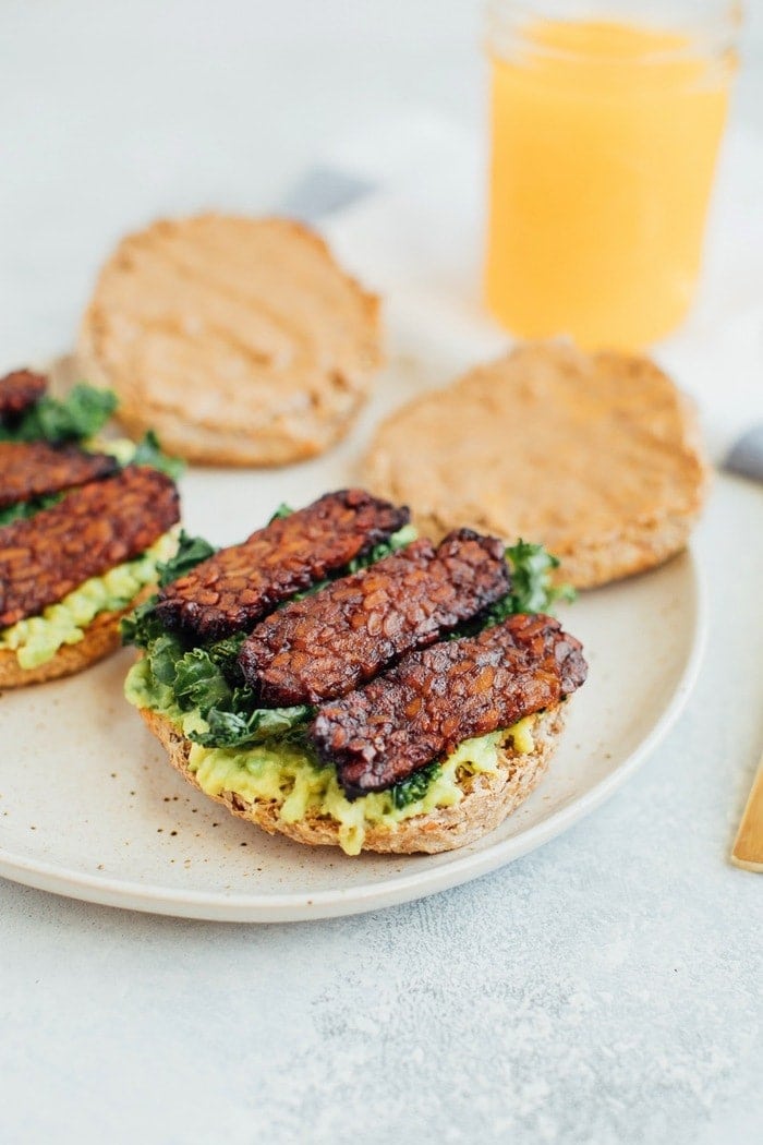 A vegan breakfast sandwich loaded with all the goods: creamy avocado, almond butter, sautéed kale and tempeh bacon, served on a perfectly toasted english muffin. A breakfast sandwich on a plate with avocado, kale, and tempeh.