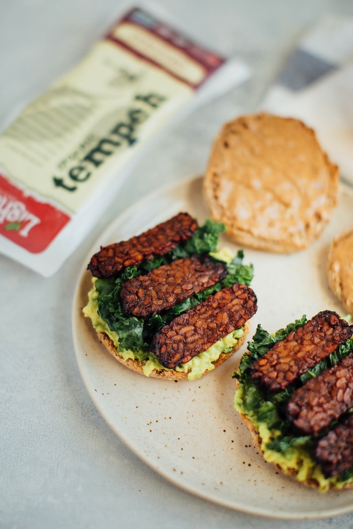 A vegan breakfast sandwich loaded with all the goods: creamy avocado, almond butter, sautéed kale and tempeh bacon, served on a perfectly toasted english muffin. A serving of a vegan breakfast sandwich on a plate with tempeh bacon, kale, avocado, and almond butter on an english muffin.