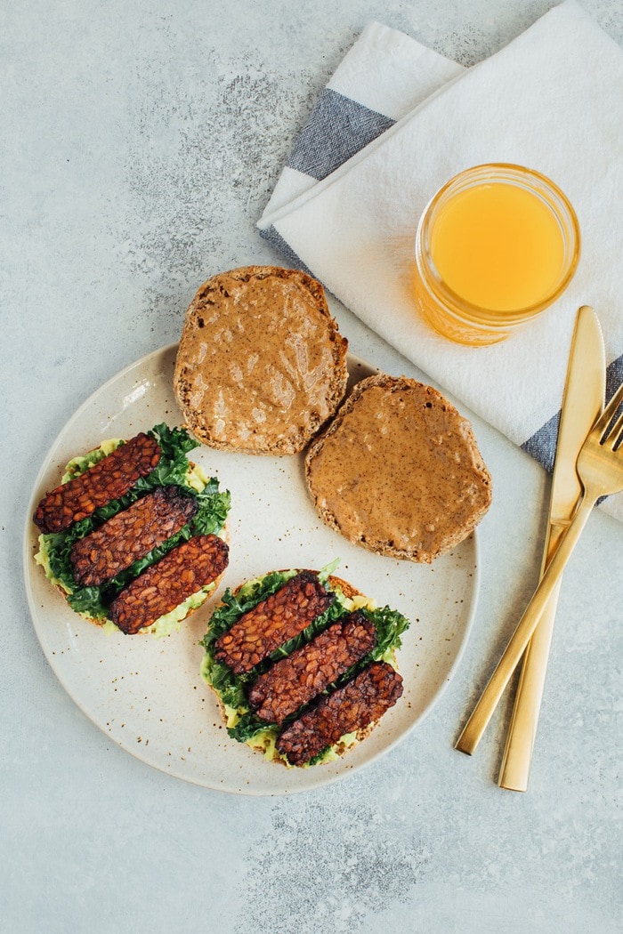 A vegan breakfast sandwich loaded with all the goods: creamy avocado, almond butter, sautéed kale and tempeh bacon, served on a perfectly toasted english muffin. A serving of a breakfast sandwich with avocado, almond butter, kale, tempeh, an english muffin, and a glass of orange juice.