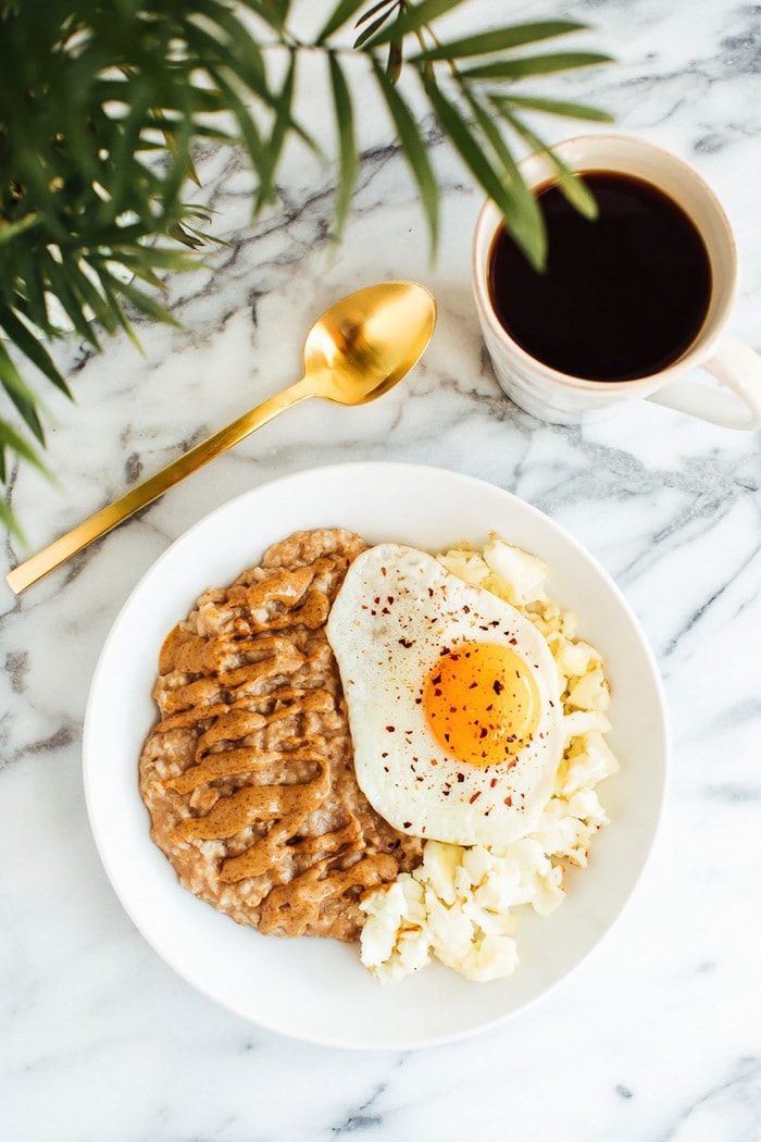 A serving of oatmeal topped with nut butter, cauliflower, and a sunny side up egg in a bowl with a cup of coffee to the side.