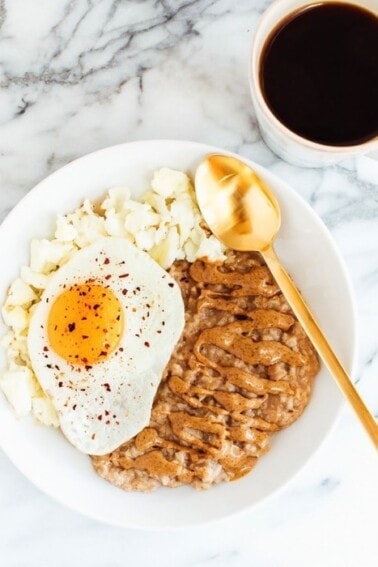 White bowl of oatmeal topped with almond butter, a fried egg with red pepper flakes, and scrambled egg whites. Black coffee next to the plate.