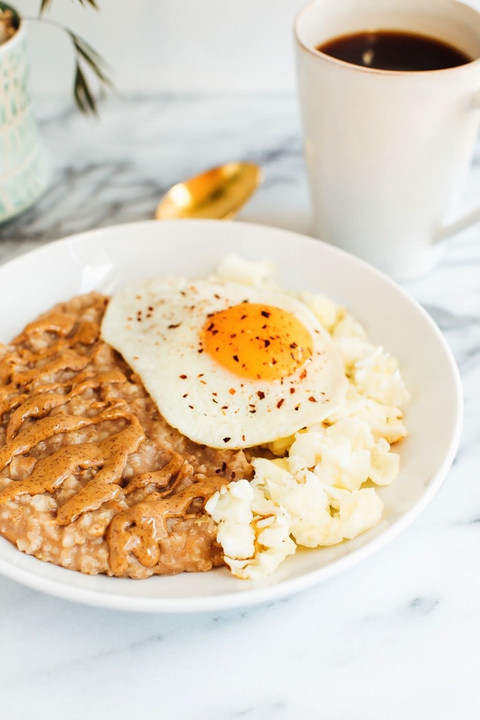 A bowl of oatmeal topped with nut butter, cauliflower, and a sunny side up egg with red pepper flakes.