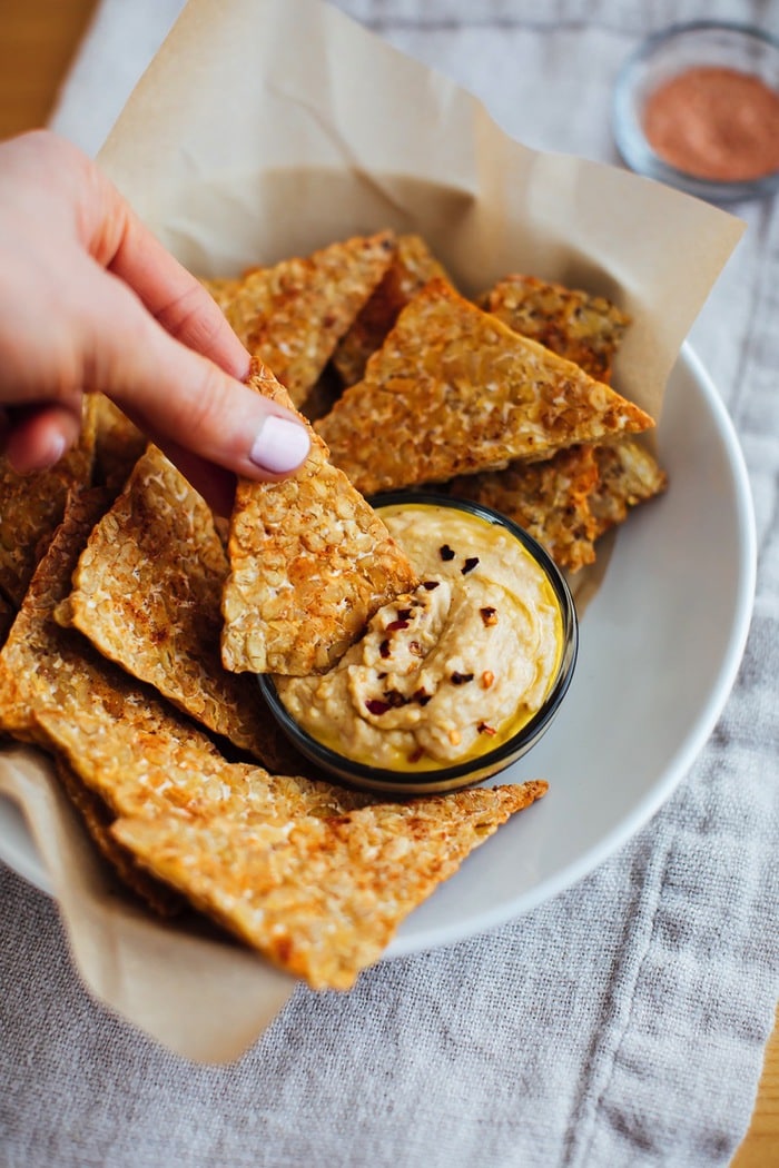 Hand with light pink nails holding a bbq baked tempeh chip and dipping it into hummus. Hand with light pink nails holding a bbq baked tempeh chip and dipping it into hummus.