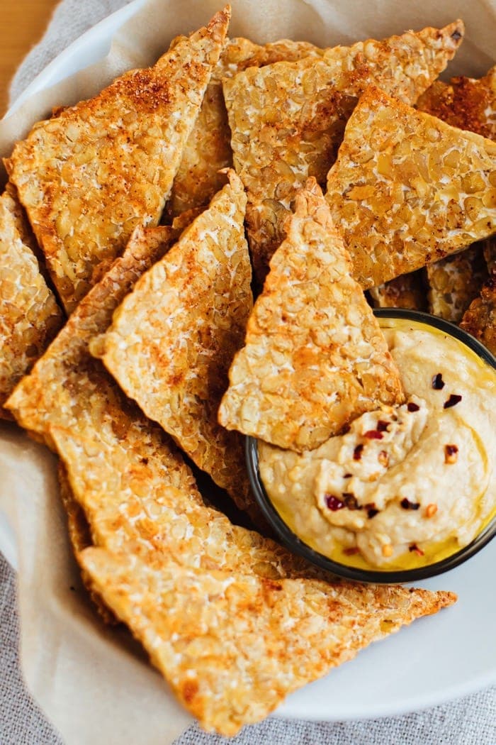 Close up of BBQ tempeh chips with hummus in a white bowl. Close up of BBQ tempeh chips with hummus in a white bowl.