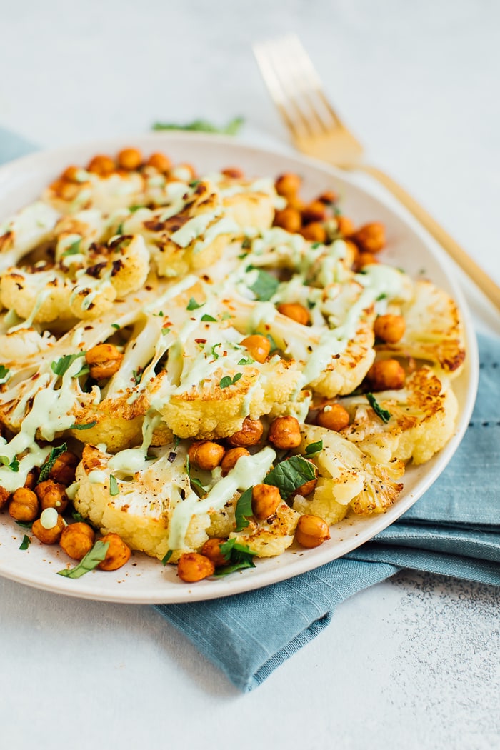 PIN THIS RECIPE for Delicious Roasted Cauliflower Steaks! Roasted cauliflower steaks on a tan plate with roasted chickpeas and a green tahini sauce.