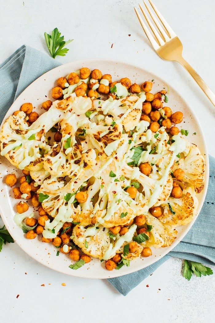PIN THIS RECIPE for Delicious Roasted Cauliflower Steaks! Cauliflower steaks with roasted chickpeas and a green tahini sauce on a plate with a gold fork in the background.