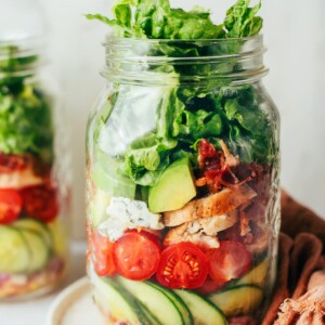A glass mason jar layered with fresh vegetables such as cucumber slices, cherry tomatoes, red onion, lettuce, avocado, and cooked chicken.