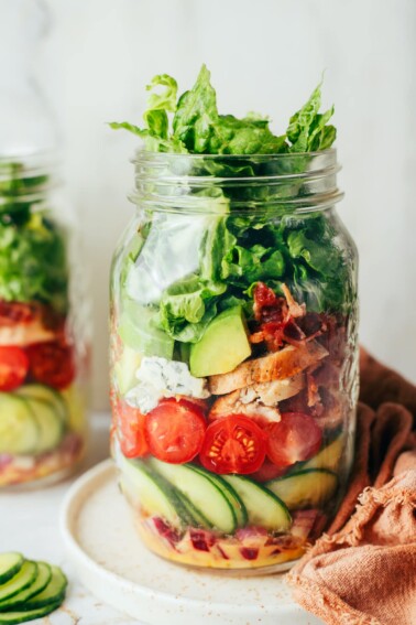 A glass mason jar layered with fresh vegetables such as cucumber slices, cherry tomatoes, red onion, lettuce, avocado, and cooked chicken.