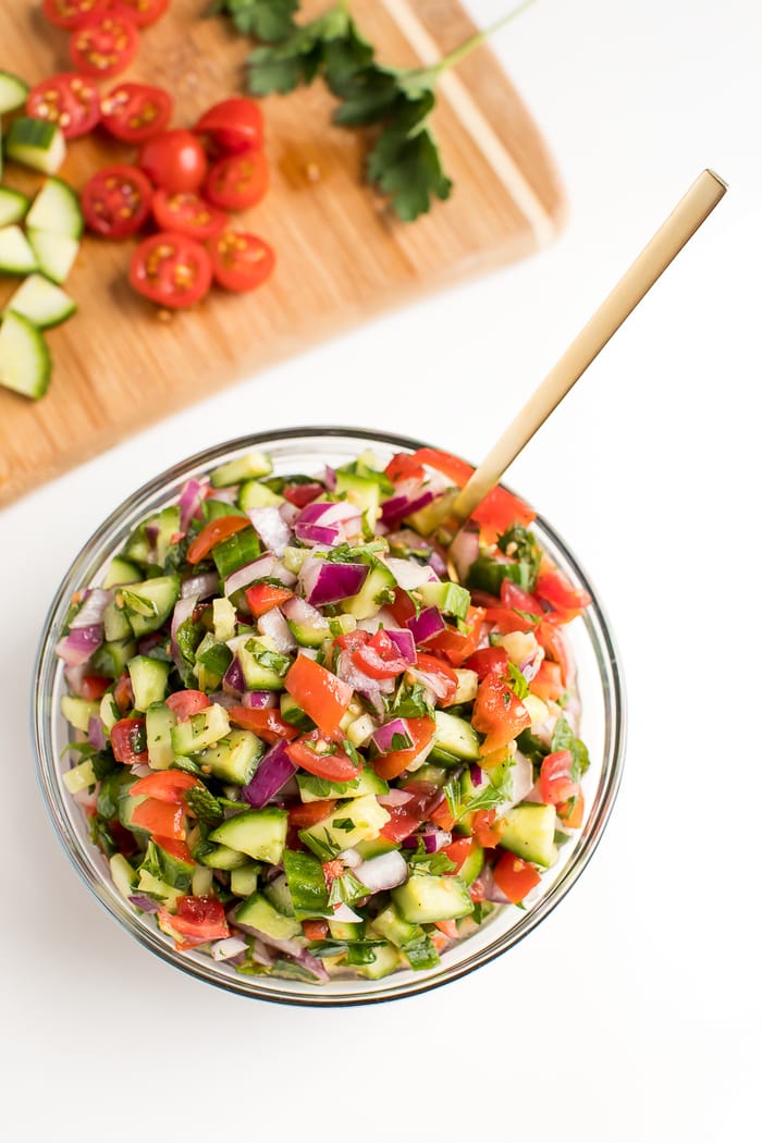PIN THIS --> Simple Israeli Salad A salad in a bowl with a large serving spoon. The salad contains tomato, cucumber, and red onion.