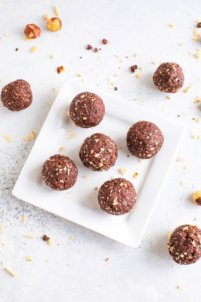 PIN THIS --> Healthy Homemade Ferrero Rocher Bites Overhead photo of homemade chocolate hazelnut energy bites on a white plate with hazelnuts around the plate.