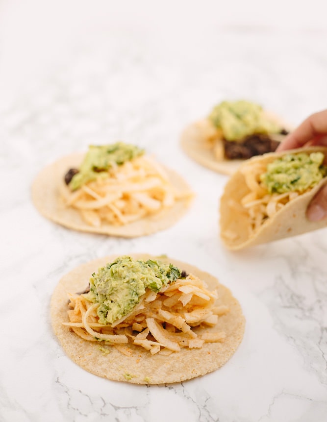 PIN THIS--> 12 Healthy Jicama Recipes Marble counter top with 4 tacos. Hand is picking up a taco. The tacos are soft tacos with black beans, jicama, and avocado mash.