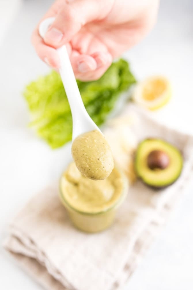 PIN THIS --> Avocado Caesar Dressing Hand holding a spoon full of avocado caesar dressing above a jar with the dressing on a table with lettuce and an avocado.