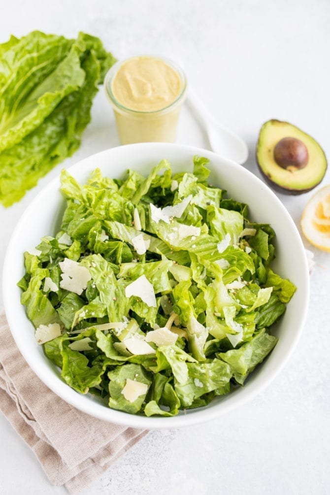 PIN THIS --> Avocado Caesar Dressing Bowl filled with romaine lettuce. Next to the bowl is a head of lettuce, a jar of avocado caesar dressing, and a half avocado.