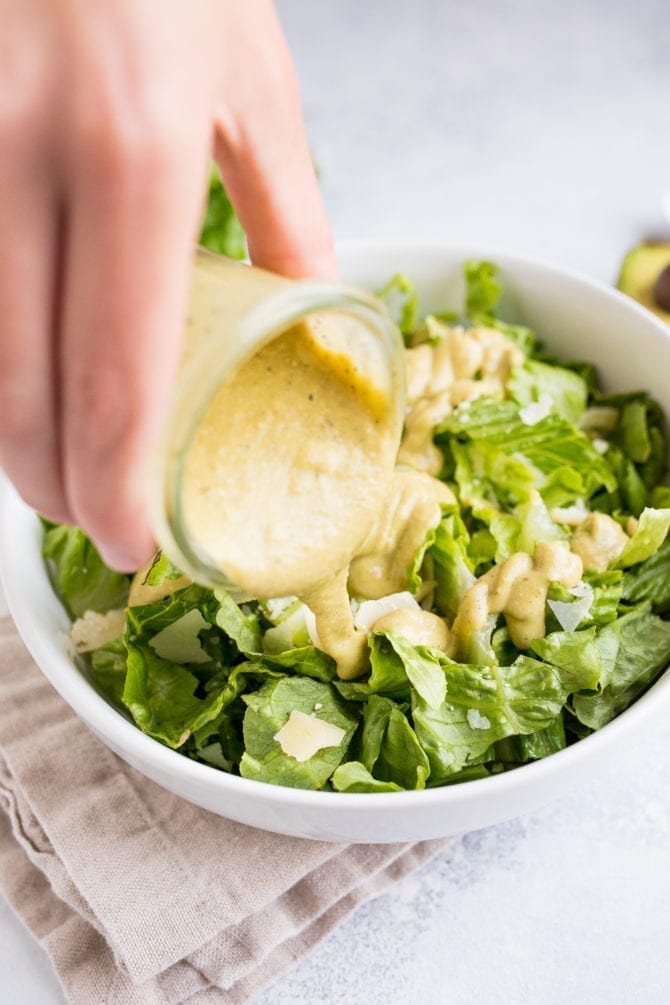 PIN THIS --> Avocado Caesar Dressing Hand pouring a jar of avocado caesar dressing over a bowl of romaine lettuce with parmesan cheese.