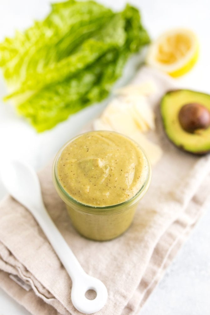 PIN THIS --> Avocado Caesar Dressing Glass jar filled with avocado caesar dressing on a table with a spoon, napkin, lettuce and avocado.