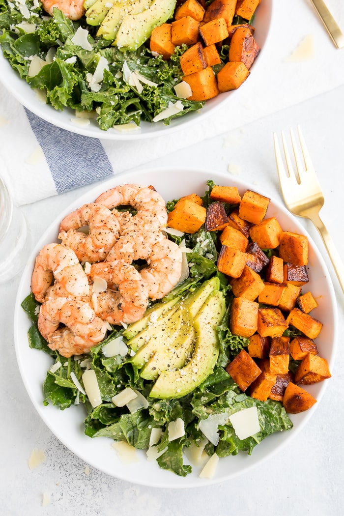 PIN THIS--> Kale Caesar Salad with Grilled Shrimp Kale caesar salad topped with grilled shrimp, avocado, sweet potatoes and parmesan cheese shavings in a white bowl with a gold fork.