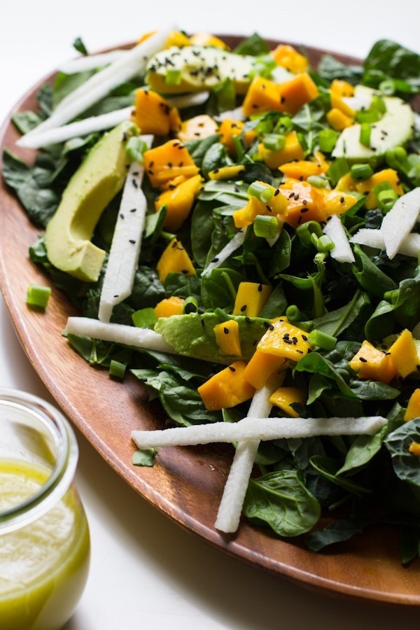 PIN THIS--> 12 Healthy Jicama Recipes Wood serving plate with a spinach, avocado, mango and jicama salad sprinkled with sesame seeds. A glass jar of lime salad dressing is on the side.