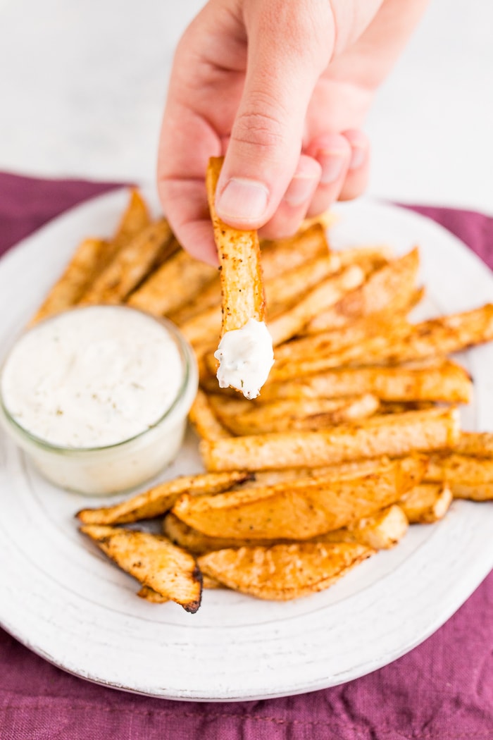 PIN THIS--> Baked Turnip Fries White plate of turnip fries over a purple napkin. A hand holds a fry that has been dipped in ranch dip.