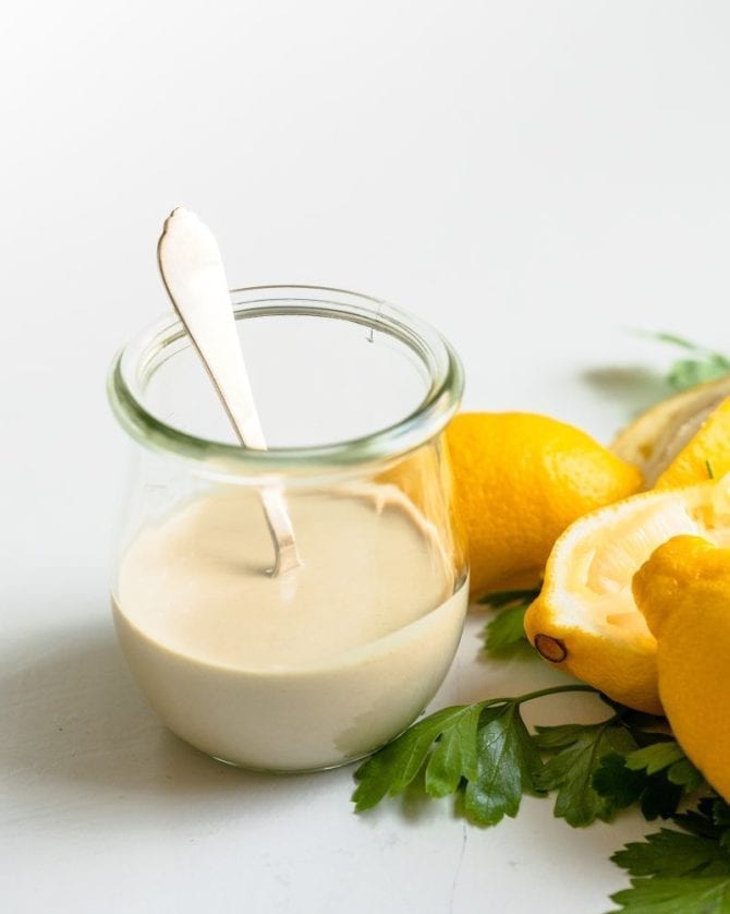 Lemon Tahini Sauce in a glass jar with a spoon in it. Lemons are next to the jar.
