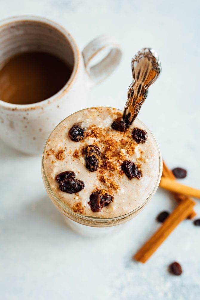 PIN THIS--> Cinnamon Raisin Overnight Oats Glass jar of cinnamon raisin overnight oats, topped with cinnamon and raisins, and a silver spoon in the jar. Cinnamon sticks, raisins, and a mug of coffee off to the side on the table.