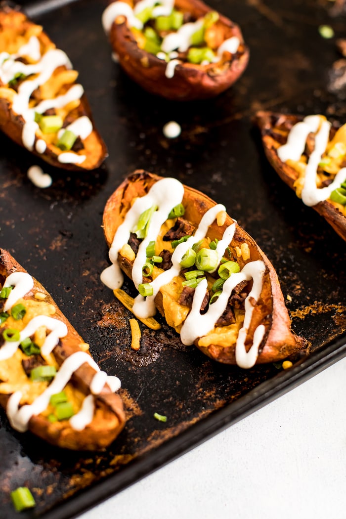 Close up photo of a sweet potato skin on a baking stone. The skins are filled with vegan cheese, bacon, chives, and sour cream drizzle.