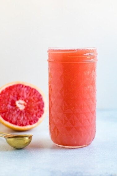A tall glass mason jar of a grapefruit drink with a half cut grapefruit in the background.