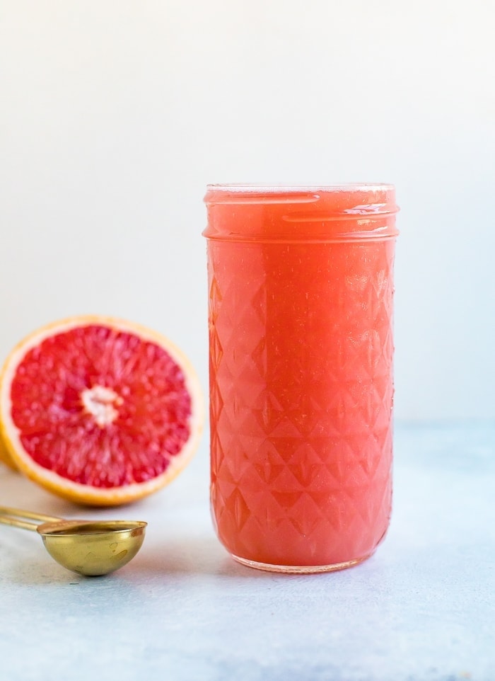 A tall glass mason jar of a grapefruit drink with a half cut grapefruit in the background.