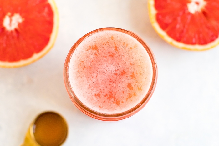 A glass jar of a grapefruit drink. Half cut grapefruit and spoon beside the jar.