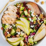 A serving of a salad topped with pears, grilled chicken, turkey bacon, cranberries, goat cheese, and walnuts.