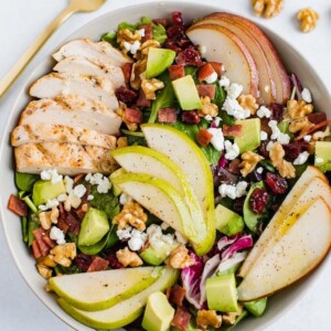 A serving of a salad topped with pears, grilled chicken, turkey bacon, cranberries, goat cheese, and walnuts.