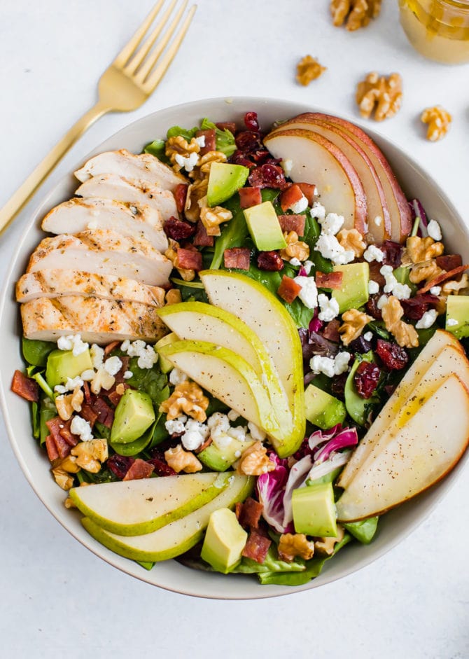 A serving of a salad topped with pears, grilled chicken, turkey bacon, cranberries, goat cheese, and walnuts.